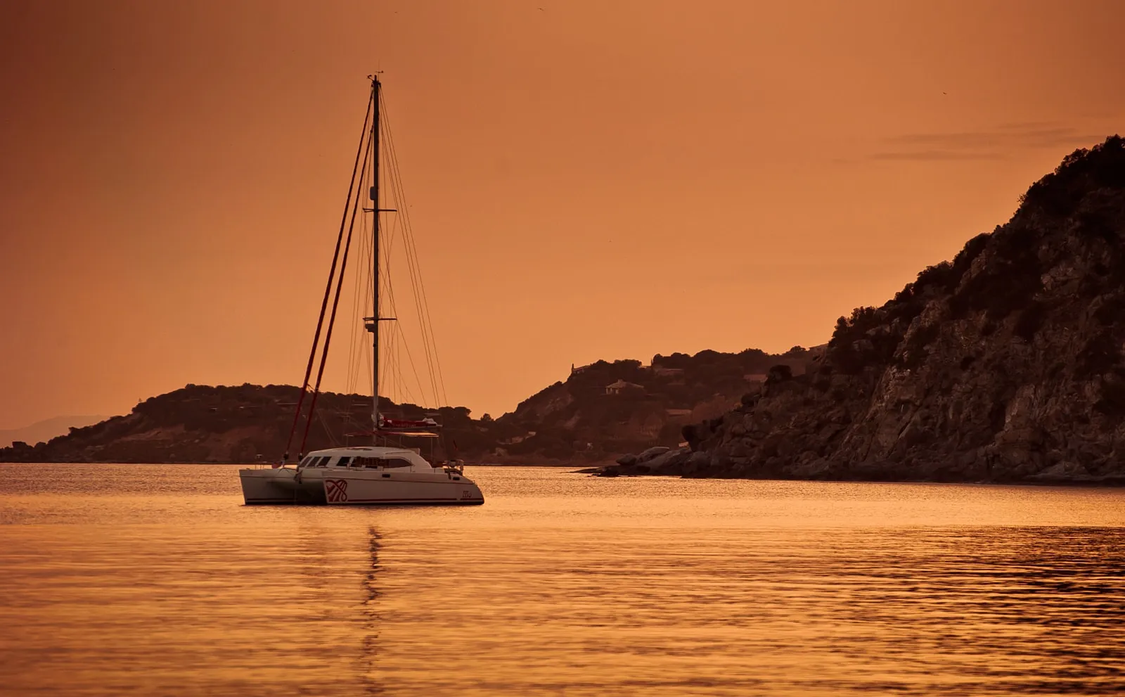 Un catamarano a vela alla fonda lungo una costa rocciosa del Mediterraneo nella luce calda del tramonto