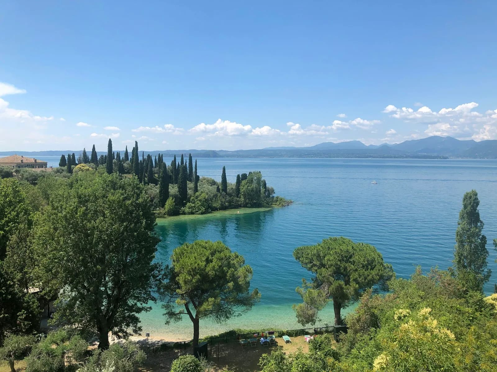 Veduta del Lago di Garda con un promontorio verde, barche e le montagne sullo sfondo