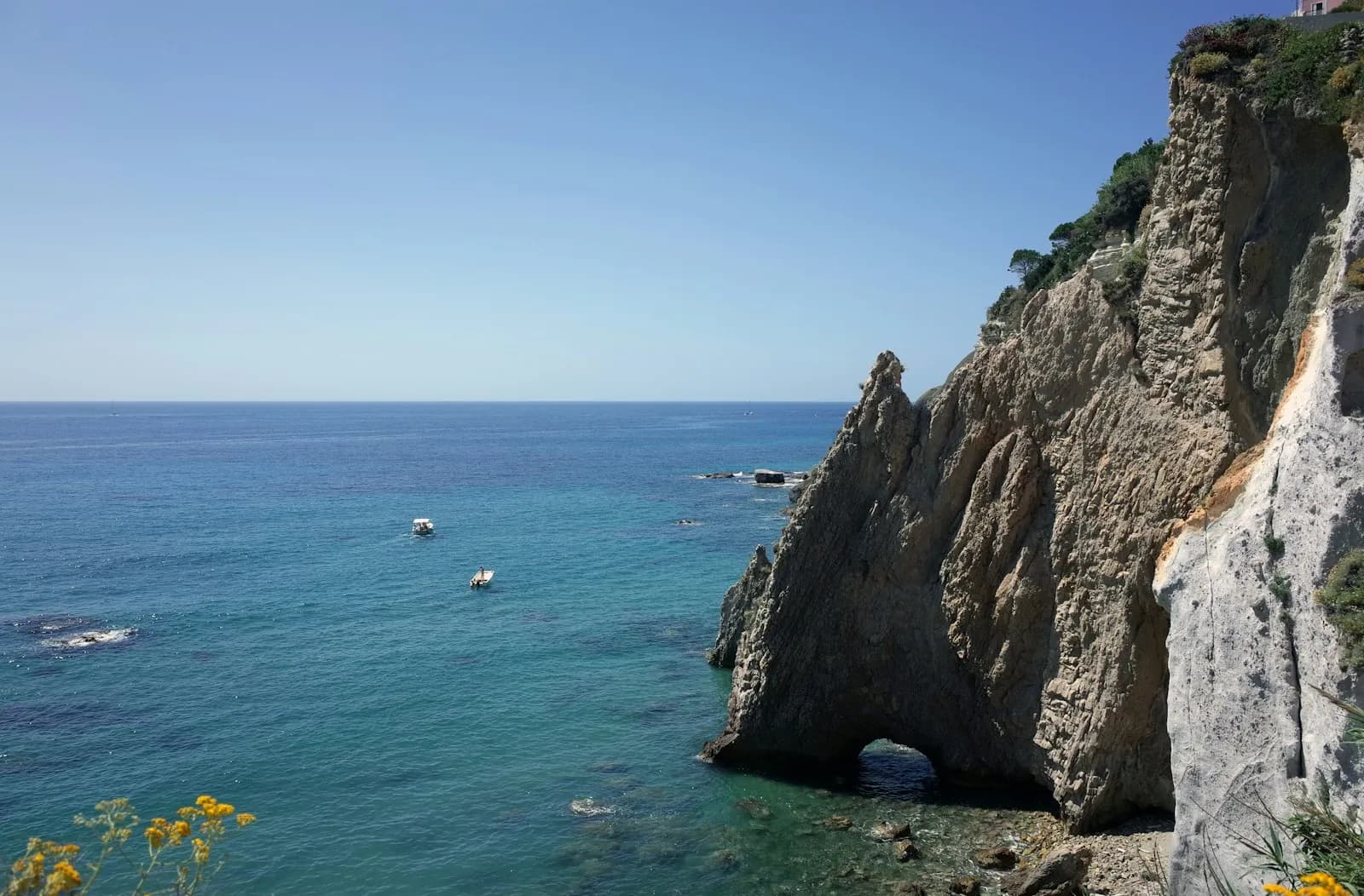Piccole barche a motore in navigazione sottocosta lungo una falesia, con il mare aperto sullo sfondo