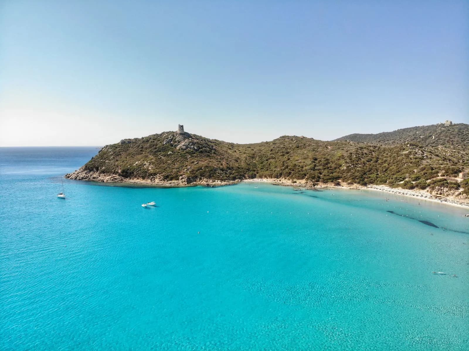 Veduta aerea della spiaggia di Porto Giunco a Villasimius, con la torre spagnola sul promontorio e barche all'ancora nell'acqua turchese