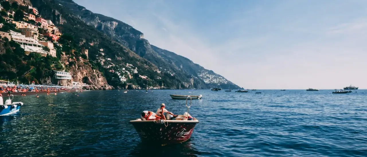 Piccola barca aperta con persone a bordo sotto una costa alta mediterranea