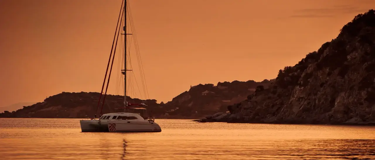 Un catamarano a vela alla fonda lungo una costa rocciosa del Mediterraneo nella luce calda del tramonto