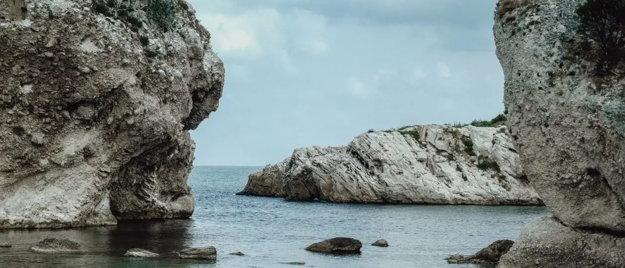 Costa rocciosa mediterranea con acqua limpida, insenatura riparata tra pareti di roccia chiara e persone in acqua