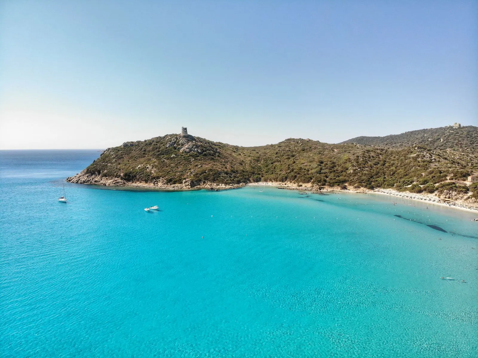 Veduta aerea della spiaggia di Porto Giunco a Villasimius, con la torre spagnola sul promontorio e barche all'ancora nell'acqua turchese